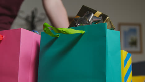 Close-Up-Of-Man-Picking-Up-Gift-Wrapped-Presents-In-Colourful-Bags-With-Tag-On-Table-In-Lounge-At-Home